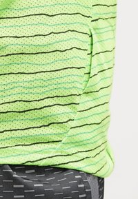 Top de tela de malla verde con patrones de rayas negras y azules. Presenta un corte holgado y un bolsillo en el lado exterior. Pantalones cortos grises con estampado debajo.