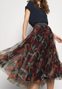 Jupe en tulle avec un motif géométrique rouge, marron et vert. Présente un design à couches et un détail scintillant à la taille.