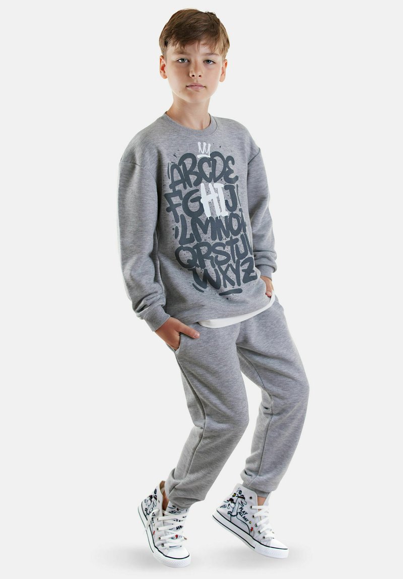 Jongen in grijze joggingbroek en trui met alfabetprint, draagt witte hoge sneakers met cartoonontwerpen, poseert met zijn handen in zijn zakken.