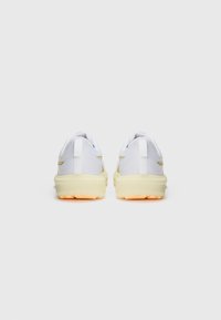 White athletic shoes with textured mesh upper, yellow accents, and a thick rubber sole featuring orange traction. Rear view displays pull tab.