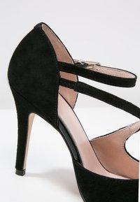 Chaussure à talon haut en daim noir avec deux brides à la cheville et une boucle, présentant un bout pointu et une doublure intérieure beige clair.