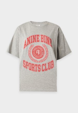 Femme aux longs cheveux noirs porte un t-shirt gris oversize avec un imprimé rouge, un jean bleu avec des fentes latérales et des talons noirs pointus, debout devant un fond blanc.