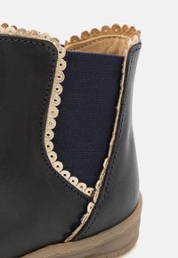 Zbliżenie czarnej skórzanej botki za kostkę z beżowym falbaniastym wykończeniem, granatową elastyczną wstawką po boku i jasnobrązową gumową podeszwą.