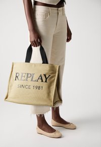 Khaki canvas torba na zakupy z czarnymi uchwytami, z wyraźnym napisem "REPLAY" i "SINCE 1981". W zestawie z jasnobeżowymi spodniami 3/4 i butami.