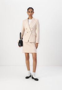 Blazer de tweed rosa claro com botões dourados, saia a condizer, mala preta, camisa branca e sapatos pretos com meias brancas. Textura suave.