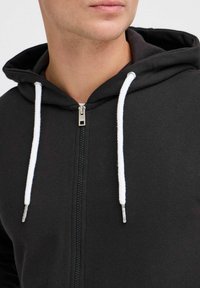 Sudadera con cremallera negra hecha de algodón, con cordones blancos, puños acanalados y una textura suave con un diseño minimalista.