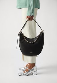 Borsa hobo in pelle nera con accentuazione in catena, tracolla regolabile e texture liscia. Indossata con pantaloni off-white e scarpe con tacco argento.