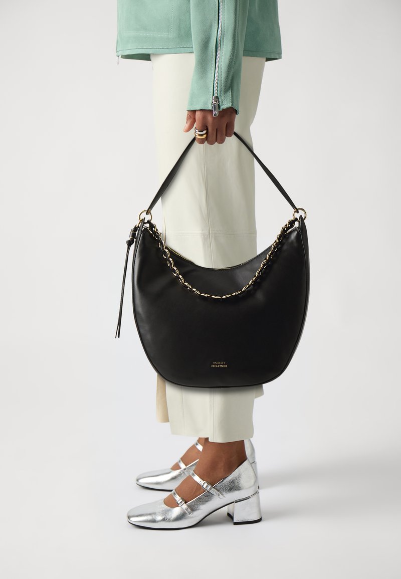 Borsa hobo in pelle nera con accentuazione in catena, tracolla regolabile e texture liscia. Indossata con pantaloni off-white e scarpe con tacco argento.