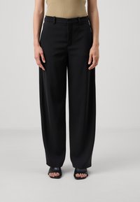 Schwarze weite Hosen aus glattem Stoff, mit hoher Taille und Gürtelschlaufen, kombiniert mit offenen schwarzen Schuhen.
