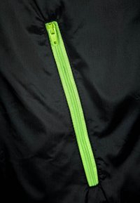 Fermuar verde strălucitor pe țesătură neagră, parțial închis, arătând cusături clare și textura materialului.