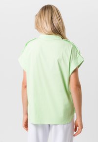 Blonde vrouw in een lichtgroen shirt met korte mouwen en opgerolde mouwen, gezien vanaf de achterkant, staande tegen een effen witte achtergrond.