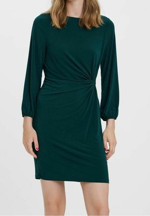 Zielona sukienka z długim rękawem z marszczeniem w talii, noszona przez osobę o średniej długości blond włosach i czerwonym lakierze do paznokci.