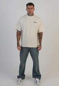 Beige oversized T-shirt med korte ærmer og rund hals. Sort logo på venstre bryst. Båret med lyseblå straight-leg jeans og hvide sneakers.