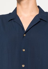 Tmavě modrá košile s krátkými rukávy a výstřihem s výřezem, zdobená světle hnědými knoflíky a hladkou, strukturovanou látkou.