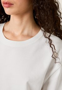 Sweatshirt gris clair à col rond, présentant une texture douce et lisse avec une coupe ample, mettant en avant un col légèrement côtelé et des cheveux bouclés à côté.