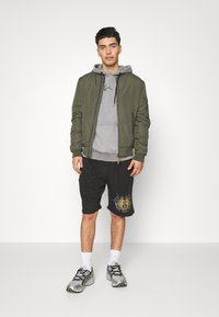 Olivgrüne Bomberjacke, grauer Hoodie darunter, schwarze Shorts mit Grafikkdruck. Graue und schwarze Sportschuhe mit gelben Akzenten.