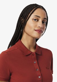 Punainen polo-paita, jossa on teksturoitu kangas, kaulus, kolme nappia ja pieni brodeerattu logo rinnassa.