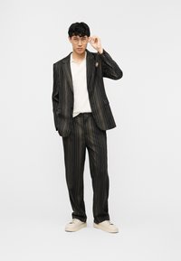 Νέος άντρας που διορθώνει τα γυαλιά του, φοράει μαύρο κοστούμι με ρίγες, λευκό πουκάμισο και μπεζ αθλητικά παπούτσια, σε απλό φωτεινό φόντο.