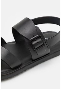 Czarne skórzane sandały z szerokimi paskami, regulowanym zapięciem i teksturowaną wkładką na czarnej gumowej podeszwie.