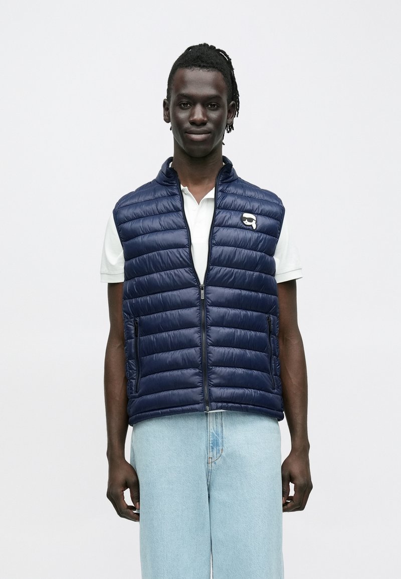 Ung mann med mørk hud og dreadlocks, iført en marineblå vattert vest over en hvit polo-skjorte og lyseblå jeans, står foran en enkel hvit bakgrunn.