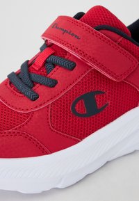 Adidași atletici roșii din plasă, cu o curea Velcro, șireturi negre și o talpă din cauciuc alb. Prezintă un logo negru pe lateral și un material texturat.