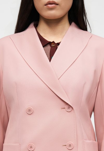 Γυναίκα που φοράει διπλόμπελο σακάκι σε ανοιχτό ροζ χρώμα πάνω από πουκάμισο μαρονού χρώματος με κουμπιά, εμφανιζόμενη από το λαιμό έως το μέσο του κορμού.