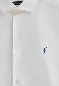 Witte blouse met een strakke kraag, voorzien van beige knopen en een kleine marineblauwe geborduurde figuur aan de linkerkant. Glad stoftextuur.