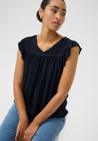 Haut bleu marine à col en V avec de courtes manches volantées et un tissu texturé ; accent en dentelle sur la poitrine ; porté avec un jean bleu clair.