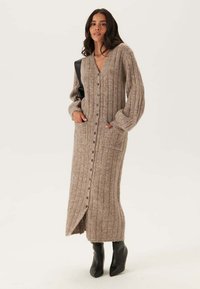 Lang, geribbelde cardigan in lichtbruin met een V-hals, knopen aan de voorkant, twee zakken en brede mouwen, gecombineerd met zwarte enkellaarsjes.