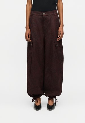Φαρδιά παντελόνια cargo σκούρου μπορντώ με τσεπάκια στο πλάι που κλείνουν με κουμπιά και ρέλι με κορδόνια, φορεμένα με μαύρα παπούτσια μυτερά στη μύτη.