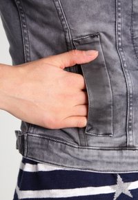 Una mano parzialmente all'interno della tasca laterale di una giacca in denim grigio, indossata sopra un indumento a righe blu navy e bianche con un motivo a stelle.