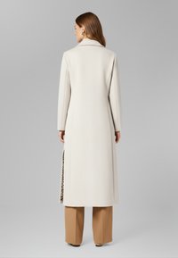 Cappotto lungo color crema con superficie strutturata e spacco laterale che rivela una fodera con stampa leopardata, indossato su pantaloni beige a gamba larga.