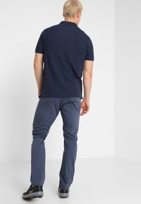 Homme aux cheveux blonds portant un polo bleu marine, un pantalon bleu et des chaussures de sport noires, debout de dos devant un fond blanc.