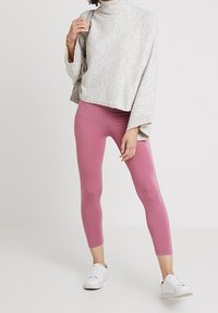 Kvinna som bär en ljusgrå polotröja, rosa åtsittande leggings, vita sneakers och bär en beige axelväska.