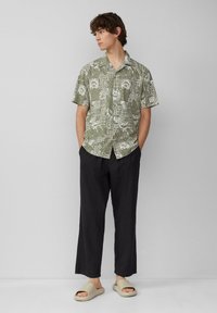 Camicetta a maniche corte verde oliva con motivi floreali e paisley bianchi, pantaloni neri larghi e sandali beige a scivolo. Design semplice e rilassato.