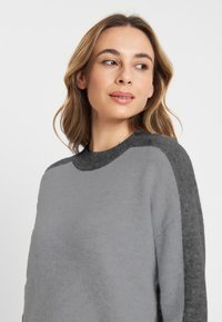 Grijze trui met een lichter grijze body en donkergrijze accenten op de schouders en kraag, met een zachte textuur en een relaxte pasvorm.