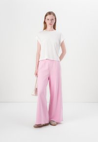 Hvid, kortærmet top kombineret med pink, brede bukser og grå slip-on sko. Modellen holder en lille, lys håndtaske.