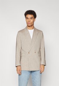 Beige linen double-breasted blazer with notch lapels, side pockets, and decorative buttons, worn over a white t-shirt and light blue jeans.