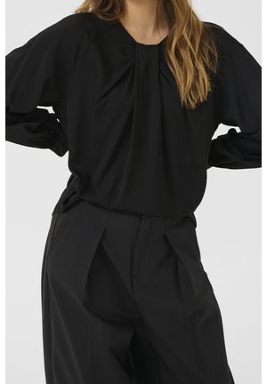 Femme portant un chemisier ample noir plissé à manches longues et un pantalon noir à large jambe taille haute, mains sur les hanches.