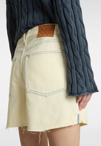 Shorts en denim de couleur jaune clair avec un ourlet effiloché, des coutures bleu contrastées et deux poches arrière. Étiquette de patch en cuir brun texturé sur la taille.