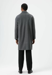 Grijze wollen jas met een getailleerde kraag, lange mouwen en een rechte silhouet, gecombineerd met een zwarte broek en schoenen. Achteraanzicht.