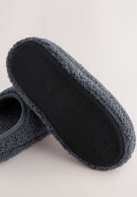 Grauer Hausschuh mit flauschiger Außenfläche, runder Form und schwarzer, strukturierter Gummisohle. Verfügt über eine glatte blaue Einfassung am Einstieg. Größe angegeben als 42.