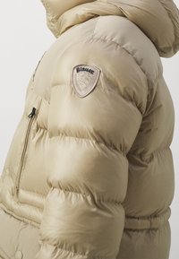 Beżowa pikowana kurtka o gładkiej, błyszczącej fakturze. Posiada kieszeń na zamek oraz naszywkę z logo na górnej części rękawa.