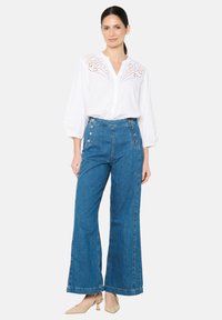 Blusa blanca de botones con detalles bordados, combinada con pantalones vaqueros de pierna ancha de cintura alta en azul, con botones laterales. Zapatos de tacón en tono neutro.