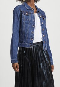 Kvinna som bär en blå denimjacka över en vit topp och en glänsande svart plisserad kjol, stående mot en enkel ljus bakgrund.