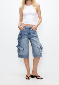 Bermudas de denim azul claro con múltiples bolsillos laterales, un corte relajado y dobladillos deshilachados, combinadas con una blusa sin mangas blanca y sandalias negras.