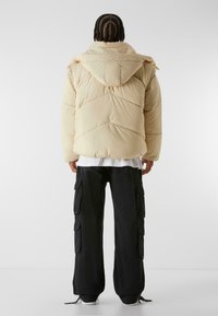 Beige puffer jacket with a hood, featuring quilted stitching and a relaxed fit. Paired with black cargo pants with side pockets.