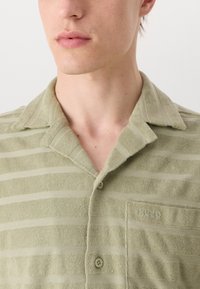 Green terry cloth shirt with horizontal stripes, a collar, button closure, and a chest pocket featuring the embossed logo "HUGO."