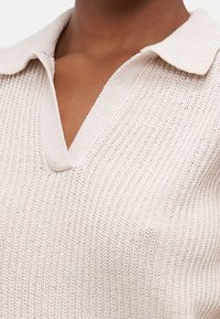 Pull en maille beige clair avec col en V et col, présentant un motif texturé et une apparence douce et confortable.