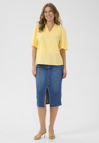 Blouse jaune à manches courtes avec un décolleté en V et des volants, associée à une jupe crayon en denim avec une fente à l'avant. Portée avec des sandales à talons hauts beige.